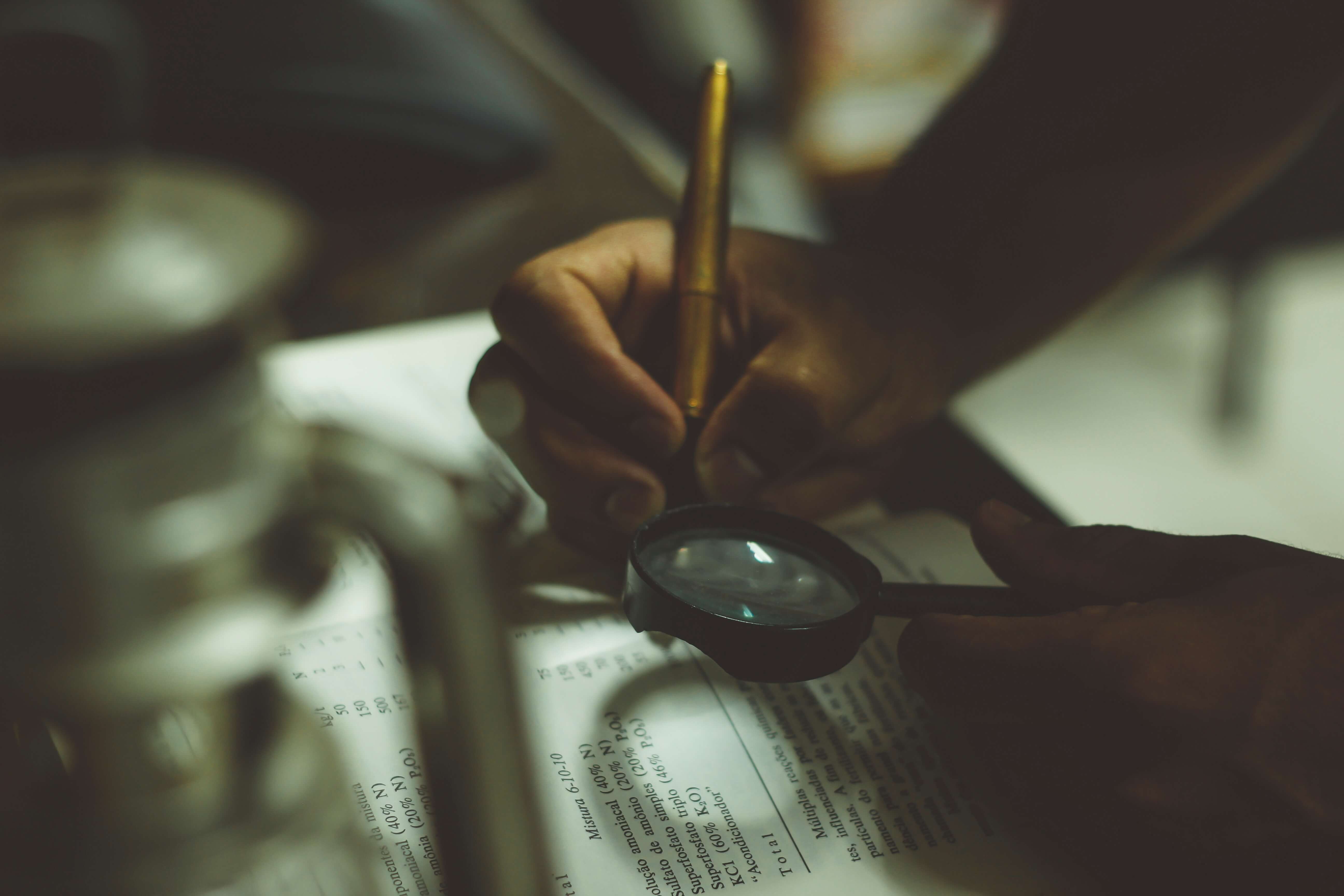 Person using a magnifying glass to read small print and a pen to make notes in a book.