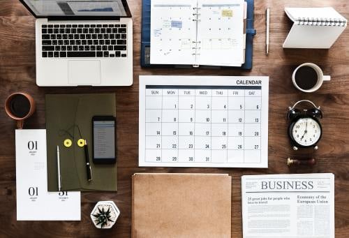 Desktop with a calendar, agenda, laptop, newspaper, cell phone, and other business items.